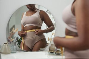 Photo of a full figured lady in front of a mirror measuring her waist with a ribbon.